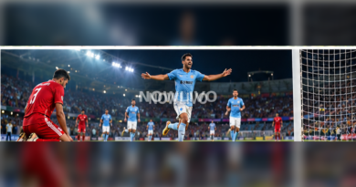 Blue-shirted footballer celebrating a goal as a red-shirted opponent looks on under stadium lights